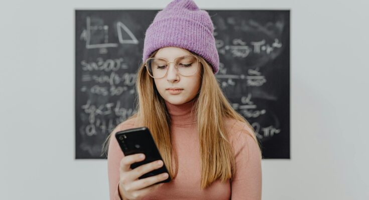 portrait of a young woman in eyeglasses in front of a blackboard using smartphone