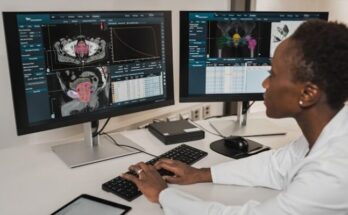 A person in a white coat typing on a keyboard at two medical monitors