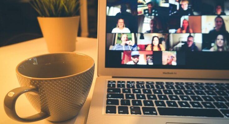 A laptop displaying a video conference with many people beside a coffee mug