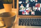 A laptop displaying a video conference with many people beside a coffee mug